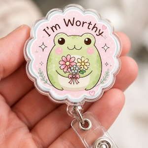 May include: A circular acrylic badge reel with a scalloped edge featuring a light green frog holding a bouquet of pink, yellow, and blue flowers. The background is light pink with sparkles and the text I'm Worthy printed in dark brown letters.