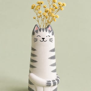 May include: A white ceramic cat-shaped vase with gray stripes and a smiling face. The vase holds a bouquet of small yellow flowers. The cat's tail wraps around the vase. A whimsical and decorative item for home decor.
