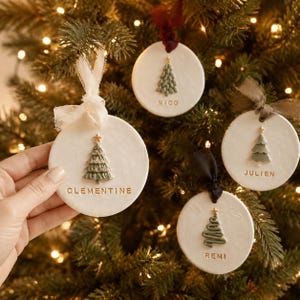 Könnte beinhalten: Vier runde, weiße Tonanhänger mit geprägten grünen Weihnachtsbäumen, goldenen Sternen und personalisierten Namen in goldener Schrift. Jeder Anhänger ist mit einem Stoffband in Weiß, Dunkelrot, Olivgrün oder Schwarz versehen.