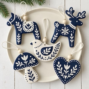 May include: Five ceramic ornaments in dark blue and white featuring a horse, a reindeer, a bird, a mushroom, and a heart. Each piece has a white string loop with a small wooden bead for hanging, decorated with intricate white floral patterns.
