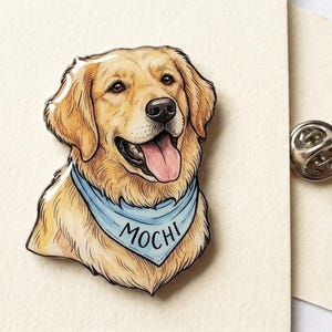 Könnte beinhalten: Eine Emaille-Anstecknadel in Form eines Golden Retrievers mit hellbraunem Fell, schwarzer Nase und geöffnetem Maul mit rosa Zunge. Der Hund trägt ein hellblaues Halstuch mit dem Namen MOCHI in schwarzen Buchstaben. Ein Metallverschluss ist sichtbar.