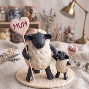 Peut inclure: Deux figurines de moutons, l'un grand et l'autre petit. Le grand mouton tient un cœur avec l'inscription "MUM", et le petit mouton tient un cœur avec "ANNA". Les moutons sont noirs et blancs.