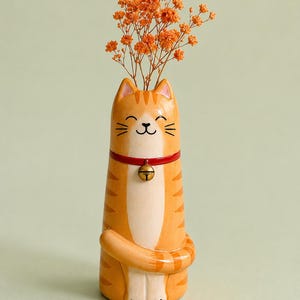 May include: A whimsical orange and white ceramic cat vase with a red collar and gold bell, holding dried orange flowers. The vase has a smiling face and a curled tail, creating a playful and decorative piece.