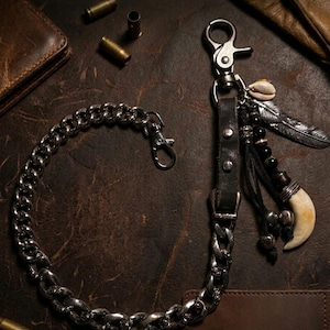 May include: A silver metal chain wallet accessory with a leather strap and decorative charms. The charms include a feather, a shell, beads, and a tooth-shaped pendant. The chain is connected to a clasp.