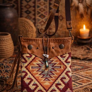 May include: A brown leather shoulder bag with a woven geometric pattern in red, yellow, and orange. The bag has a leather strap and decorative metal studs. A small pendant with feathers hangs from the flap.