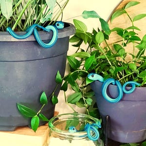 Könnte beinhalten: Dekorative blaue und türkisfarbene Schlangenfiguren, die sich um Topfpflanzen winden. Die Schlangen haben detaillierte Augen und sind in verschiedenen Töpfen und einem Glas platziert, was dem Grün eine skurrile Note verleiht. Die Töpfe sind dunkelblau.