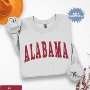 May include: A light gray sweatshirt with the word "ALABAMA" in distressed red lettering. The sweatshirt has a crew neck and long sleeves. The words "FAST SHIPPING" are in a blue burst graphic.