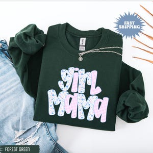 May include: Forest green sweatshirt with the text "Girl Mama" in a retro font. The letters are decorated with a pink gingham pattern and blue floral print. A silver necklace with a pendant is visible.