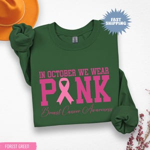 May include: Forest green sweatshirt with the text "IN OCTOBER WE WEAR PINK" in pink, along with a pink ribbon graphic and "Breast Cancer Awareness" in a script font. The sweatshirt has long sleeves.