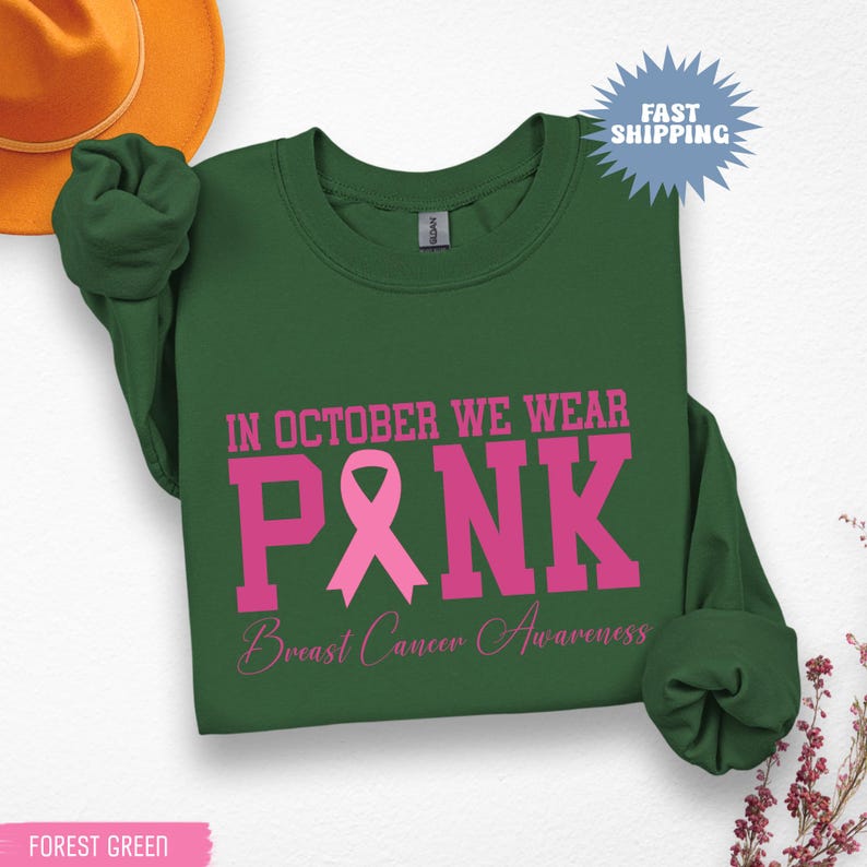 May include: Forest green sweatshirt with the text "IN OCTOBER WE WEAR PINK" in pink, along with a pink ribbon graphic and "Breast Cancer Awareness" in a script font. The sweatshirt has long sleeves.