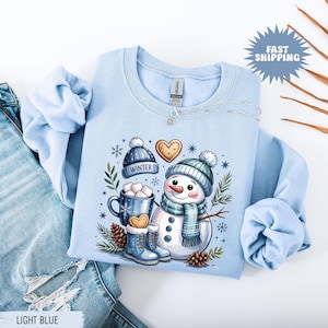 May include: Light blue sweatshirt featuring a snowman graphic with a blue hat, scarf, and boots. The design includes a mug of hot cocoa, a heart-shaped cookie, and the word "WINTER" on a hat. The sweatshirt is displayed with a pair of blue jeans.