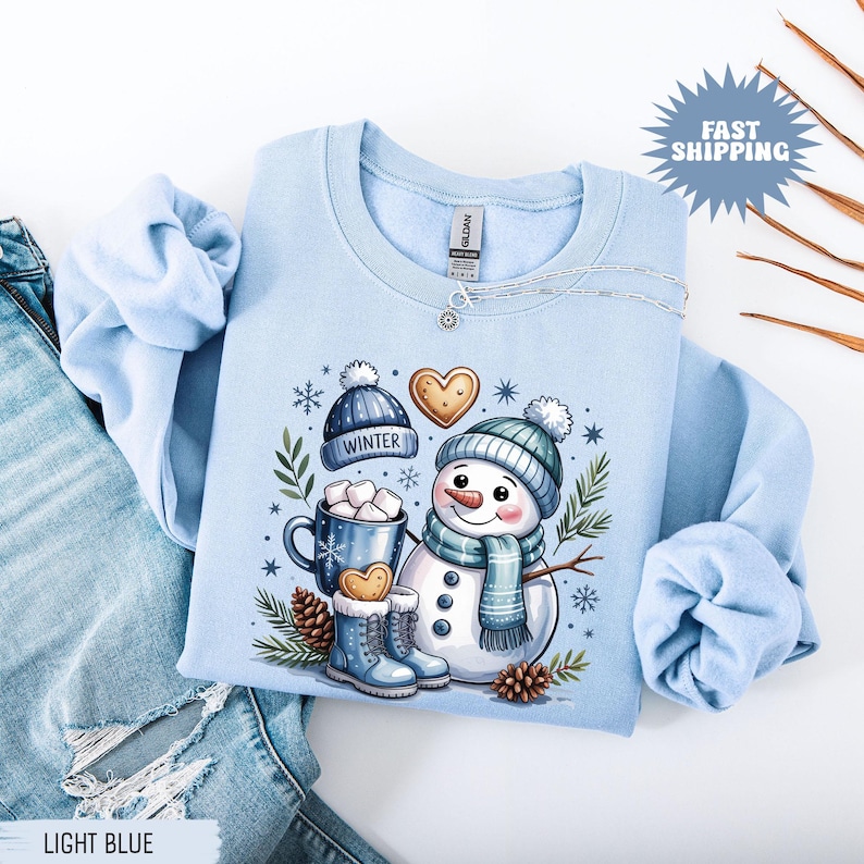 May include: Light blue sweatshirt featuring a snowman graphic with a blue hat, scarf, and boots. The design includes a mug of hot cocoa, a heart-shaped cookie, and the word "WINTER" on a hat. The sweatshirt is displayed with a pair of blue jeans.