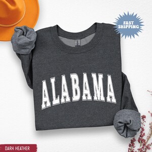 May include: Dark heather gray sweatshirt with the word "ALABAMA" in white, arched lettering. The long-sleeved shirt has a crew neck and rolled-up sleeves. A brown hat is visible in the upper left corner. The words "Fast Shipping" are in a blue burst.