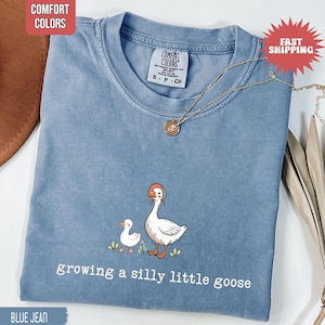 May include: Blue jean Comfort Colors t-shirt with a graphic of a goose and gosling, and the text "growing a silly little goose". A gold necklace with a pendant is draped across the shirt.