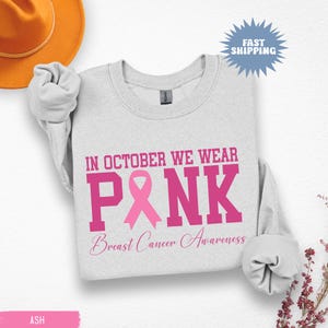 May include: Ash gray sweatshirt with the text "IN OCTOBER WE WEAR PINK Breast Cancer Awareness" in pink. A pink ribbon graphic is featured within the word "PINK". A Fast Shipping graphic is in the upper right corner.
