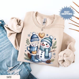 May include: Beige sweatshirt featuring a whimsical snowman design. The illustration includes a snowman wearing a blue hat and scarf, a mug of hot cocoa, and winter-themed accessories. The word "WINTER" is printed on a blue hat. The sweatshirt is folded on top of blue jeans.