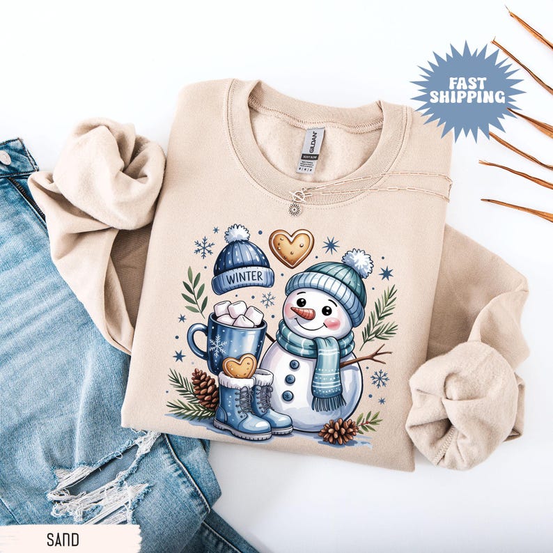May include: Beige sweatshirt featuring a whimsical snowman design. The illustration includes a snowman wearing a blue hat and scarf, a mug of hot cocoa, and winter-themed accessories. The word "WINTER" is printed on a blue hat. The sweatshirt is folded on top of blue jeans.