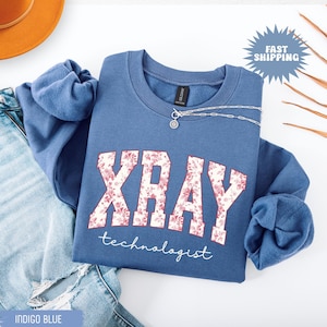 May include: Indigo blue sweatshirt with the word "XRAY" in a floral design, and the word "technologist" below. A silver necklace with a pendant is on the sweatshirt. The words "Fast Shipping" are in a blue burst.