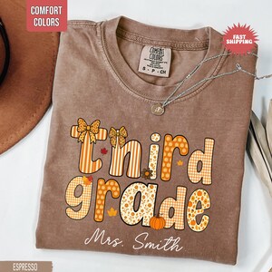 May include: Espresso-colored t-shirt with the words "Third Grade" in orange and white patterned letters. The shirt features bow accents and fall-themed graphics. The name "Mrs. Smith" is printed below the text. A silver necklace with a pendant is draped on the shirt.
