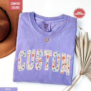 K&ouml;nnte beinhalten: Ein violettes Comfort Colors T-Shirt mit dem Wort "CUSTOM" in einem floralen Design. Das Shirt hat einen Rundhalsausschnitt und kurze &Auml;rmel. Eine goldene Halskette mit Anh&auml;nger ist auf dem Shirt drapiert. Das Shirt ist auf einer wei&szlig;en Oberfl&auml;che gefaltet.