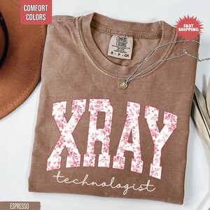 May include: Espresso-colored t-shirt with the words "XRAY" in white block letters filled with a pink floral pattern. The word "technologist" is written below in a script font. A silver necklace with a small pendant is draped on the shirt.