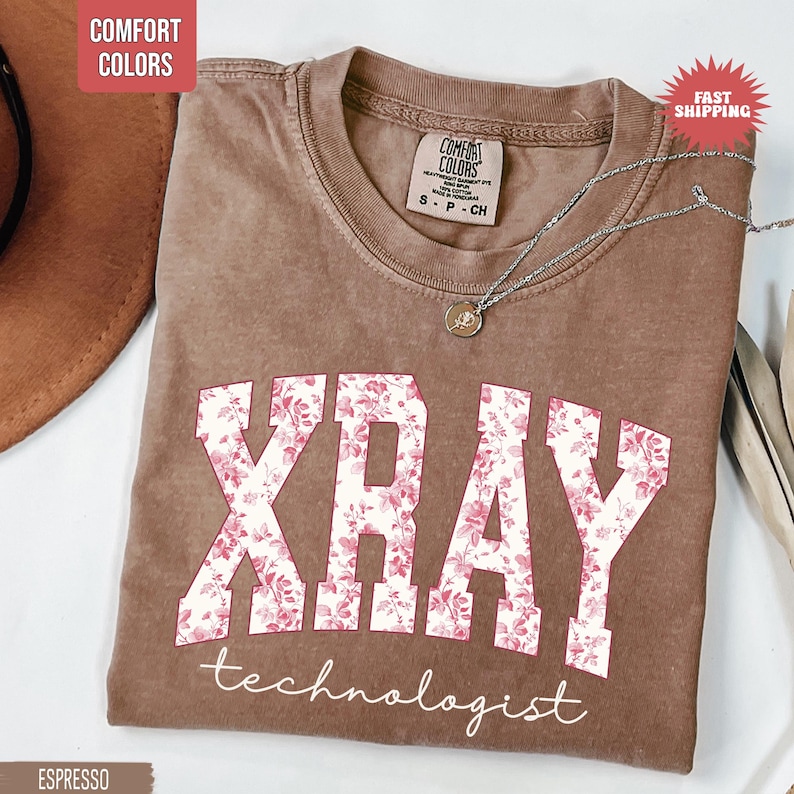 May include: Espresso-colored t-shirt with the words "XRAY" in white block letters filled with a pink floral pattern. The word "technologist" is written below in a script font. A silver necklace with a small pendant is draped on the shirt.
