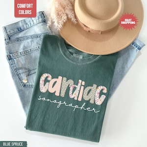 Puede incluir: Una camiseta azul abeto con la palabra "Cardiac sonographer" en un diseño floral y a rayas. La camiseta está colocada sobre un par de vaqueros azul claro, con un sombrero color beige y un collar dorado. También se ven las palabras "Comfort Colors" y "Fast Shipping".