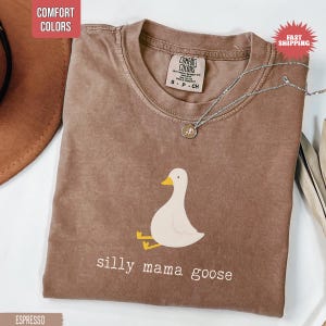 May include: Espresso-colored t-shirt with a crew neck and a graphic of a cartoon goose with the text "silly mama goose". The shirt has a vintage look and feel. A silver necklace with a pendant is on the shirt.