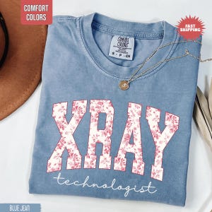 May include: Blue jean Comfort Colors t-shirt with "XRAY" in white block letters filled with a pink floral pattern. The word "technologist" is written in white script below the letters. A gold necklace with a pendant rests on the shirt.