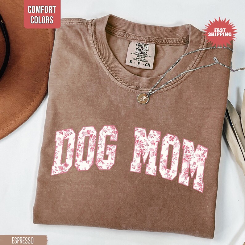 May include: Espresso-colored Comfort Colors t-shirt with the words "DOG MOM" in a white font filled with a pink floral pattern. A silver necklace with a pendant is draped across the shirt.