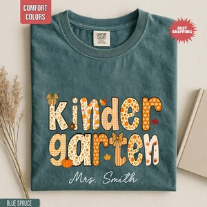 May include: Blue spruce t-shirt with the word "Kindergarten" in orange and white fall-themed lettering. The shirt includes the name "Mrs. Smith" in white script. The shirt has a Comfort Colors tag and a "Fast Shipping" sticker.