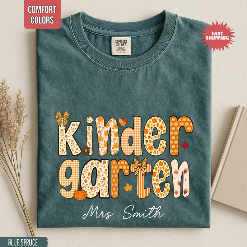 May include: Blue spruce t-shirt with the word "Kindergarten" in orange and white fall-themed lettering. The shirt includes the name "Mrs. Smith" in white script. The shirt has a Comfort Colors tag and a "Fast Shipping" sticker.