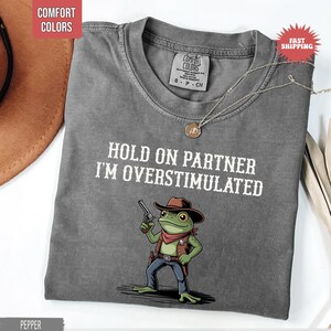 May include: Pepper-colored t-shirt with the text "HOLD ON PARTNER I'M OVERSTIMULATED" and a cartoon frog wearing a cowboy hat and holding a gun. The shirt has a crew neck and short sleeves. A silver necklace with a pendant is on the shirt.