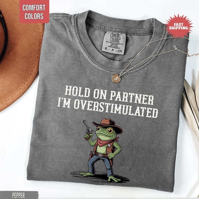 May include: Pepper-colored t-shirt with the text "HOLD ON PARTNER I'M OVERSTIMULATED" and a cartoon frog wearing a cowboy hat and holding a gun. The shirt has a crew neck and short sleeves. A silver necklace with a pendant is on the shirt.