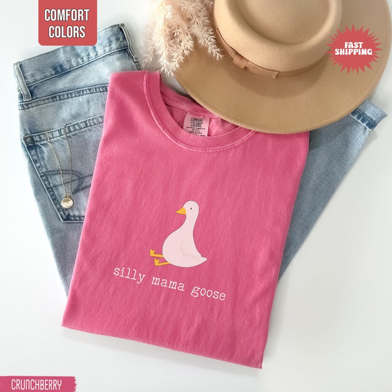 May include: A pink t-shirt with a graphic of a cartoon goose and the text "silly mama goose". The shirt is folded on top of a pair of blue jeans, with a tan hat and dried flowers in the background. The shirt is labeled "Comfort Colors".