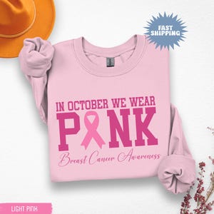 May include: Light pink sweatshirt with the text "IN OCTOBER WE WEAR PINK Breast Cancer Awareness" in a bold, dark pink font. A pink ribbon graphic is featured within the word "PINK". A light orange hat is visible in the upper left corner.