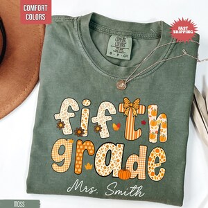 May include: A moss green Comfort Colors t-shirt with the words "Fifth Grade" in a fall-themed design. The design includes floral patterns, pumpkins, and the name "Mrs. Smith" at the bottom. A silver necklace with a pendant is draped across the shirt.
