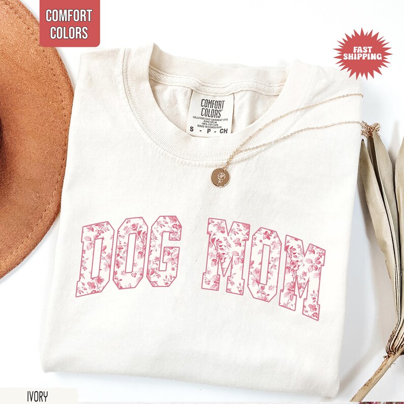 May include: Ivory-colored t-shirt with the text "DOG MOM" in a pink floral pattern. The shirt has a crew neck and short sleeves. A gold necklace with a pendant is draped on the shirt.