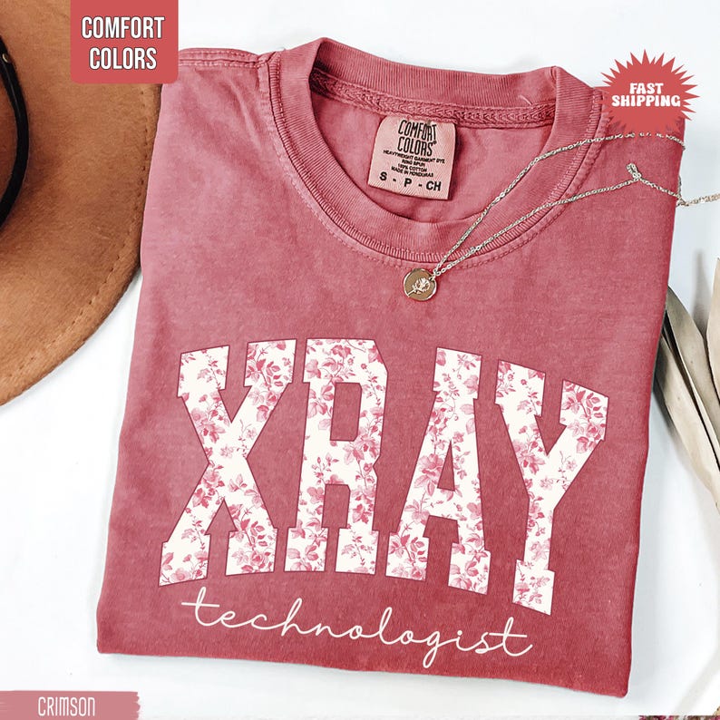 May include: Crimson-colored t-shirt with the words "XRAY technologist" in white, floral-filled letters. A silver necklace with a pendant rests on the shirt. The shirt has a crew neck and a tag that says "Comfort Colors."
