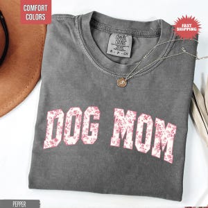 May include: A gray t-shirt with the text "DOG MOM" in a floral pattern. The shirt has a crew neck and short sleeves. A silver necklace with a pendant is draped on the shirt.