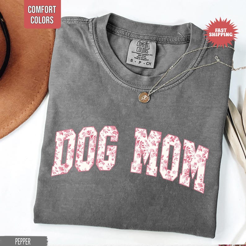 May include: A gray t-shirt with the text "DOG MOM" in a floral pattern. The shirt has a crew neck and short sleeves. A silver necklace with a pendant is draped on the shirt.