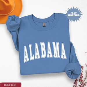 May include: A blue crewneck sweatshirt with the word "ALABAMA" in white, distressed lettering. The sweatshirt has long sleeves and a ribbed neckline. The words "Fast Shipping" are in a starburst graphic. The bottom left corner has the words "Indigo Blue".