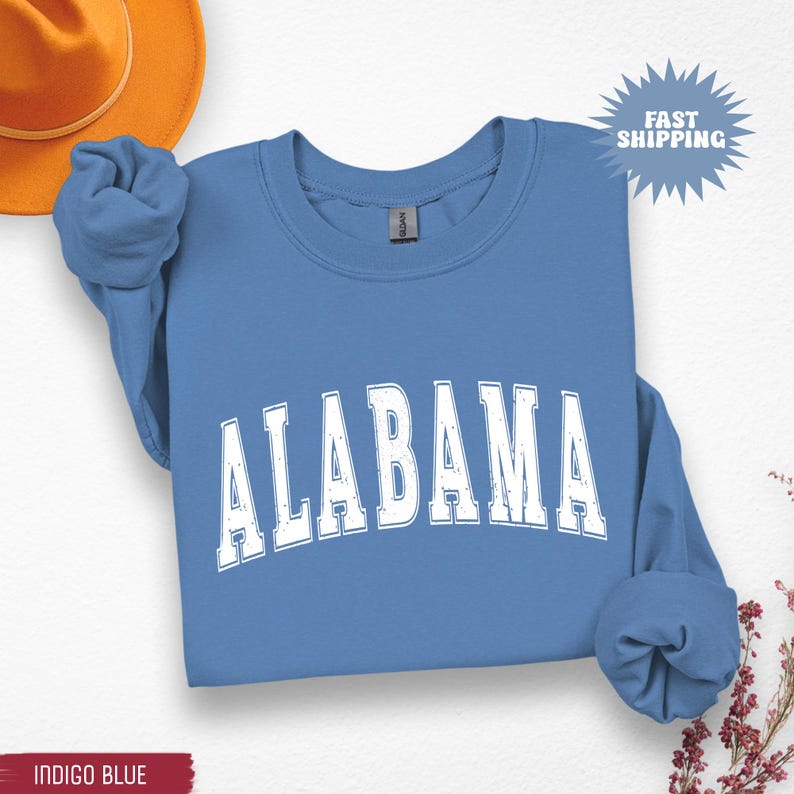 May include: A blue crewneck sweatshirt with the word "ALABAMA" in white, distressed lettering. The sweatshirt has long sleeves and a ribbed neckline. The words "Fast Shipping" are in a starburst graphic. The bottom left corner has the words "Indigo Blue".