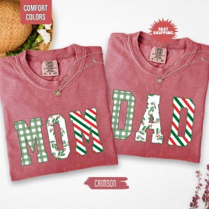 May include: Two crimson-colored t-shirts with the words "MOM" and "DAD" in large, decorative letters. The letters have green plaid, red and white striped, and floral patterns. The shirts have a crew neck and short sleeves.