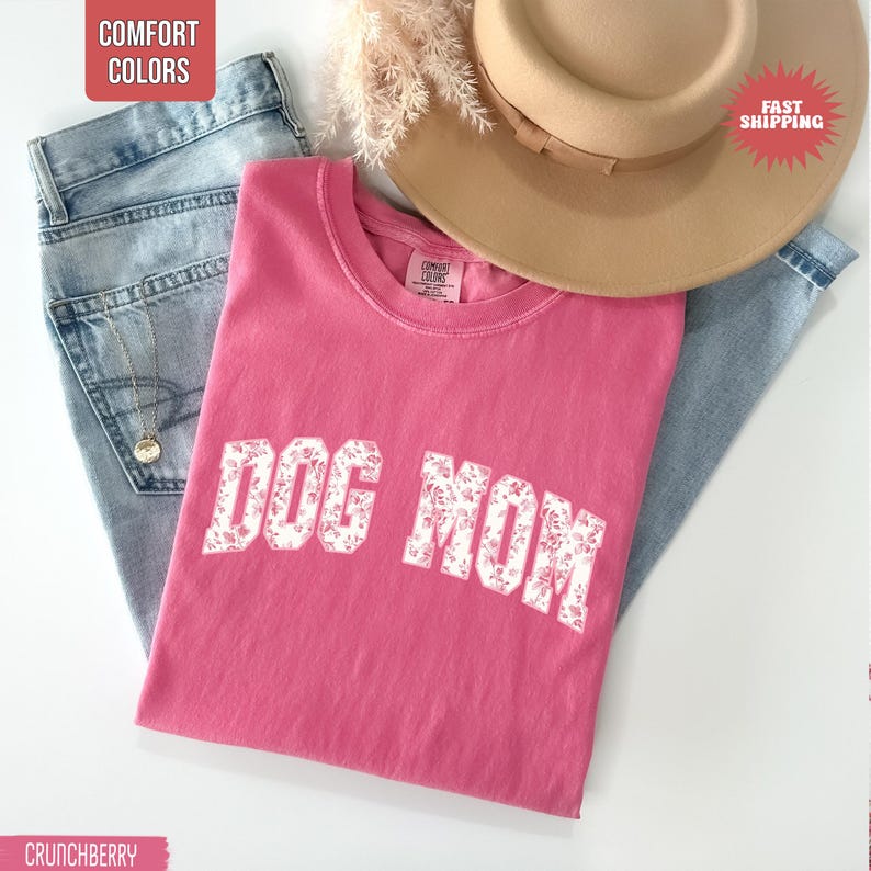 May include: A pink t-shirt with the text "DOG MOM" in a floral design, paired with light blue jeans and a tan hat. The shirt is a Comfort Colors brand.