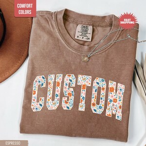 K&ouml;nnte beinhalten: Espressofarbenes T-Shirt mit dem Wort "CUSTOM" in einem floralen Design. Das Shirt hat einen Rundhalsausschnitt und kurze &Auml;rmel. Eine silberne Halskette mit Anh&auml;nger ist auf dem Shirt drapiert.