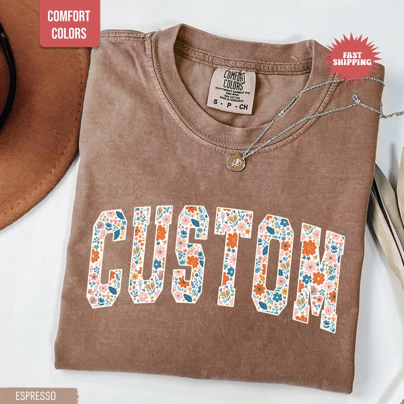 K&ouml;nnte beinhalten: Espressofarbenes T-Shirt mit dem Wort "CUSTOM" in einem floralen Design. Das Shirt hat einen Rundhalsausschnitt und kurze &Auml;rmel. Eine silberne Halskette mit Anh&auml;nger ist auf dem Shirt drapiert.