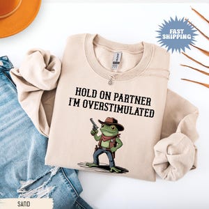 Może przedstawiać: Beżowa bluza z napisem "HOLD ON PARTNER I'M OVERSTIMULATED" i kreskówkową żabą w kapeluszu kowbojskim i z pistoletem. Bluza jest złożona na białej powierzchni z parą niebieskich dżinsów.