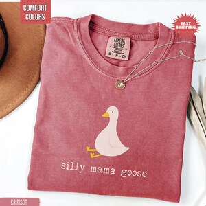 May include: Crimson Comfort Colors t-shirt with a graphic of a light pink goose with yellow feet and beak. The text "silly mama goose" is below the goose. A silver necklace with a pendant is on the shirt.