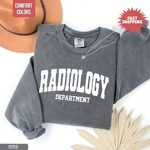 May include: A gray sweatshirt with the words "RADIOLOGY DEPARTMENT" in white block letters. The sweatshirt is a Comfort Colors brand. A gold necklace with a pendant is draped on the sweatshirt. A brown hat and a dried palm leaf are in the background.